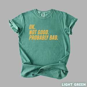 Pu&ograve; includere: T-shirt verde chiaro con la scritta gialla "OK. NOT GOOD. PROBABLY BAD." La maglietta &egrave; piegata e ha le maniche arrotolate. Le parole "LIGHT GREEN" sono in basso a destra.