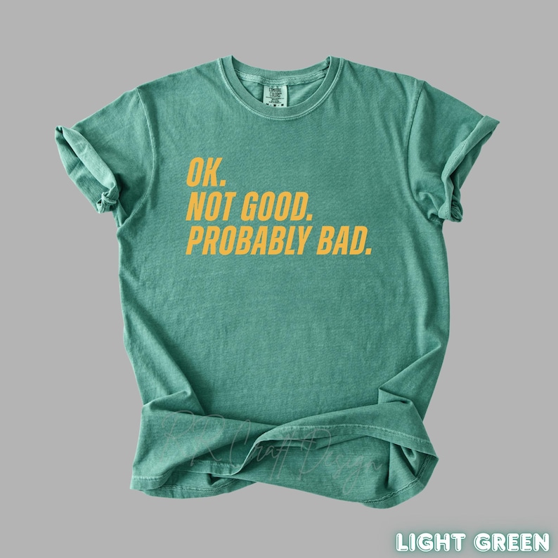Pu&ograve; includere: T-shirt verde chiaro con la scritta gialla "OK. NOT GOOD. PROBABLY BAD." La maglietta &egrave; piegata e ha le maniche arrotolate. Le parole "LIGHT GREEN" sono in basso a destra.