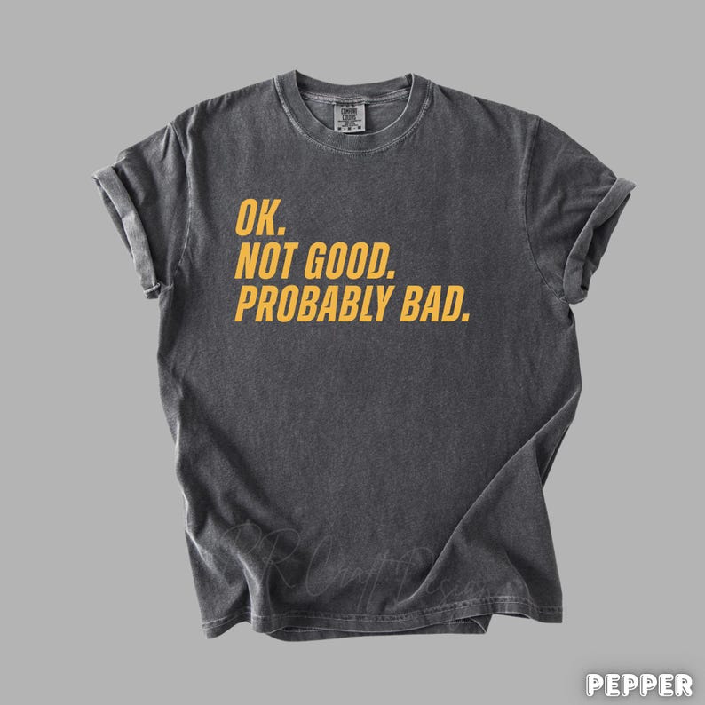 Pu&ograve; includere: T-shirt grigio scuro con il testo giallo "OK. NOT GOOD. PROBABLY BAD.". La maglietta ha un girocollo e maniche corte. Il tessuto sembra prelavato per un aspetto vintage.