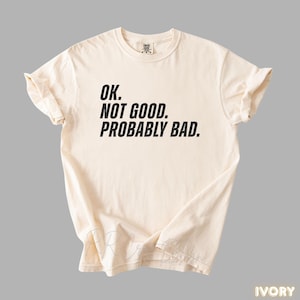 Pu&ograve; includere: T-shirt color avorio con la scritta "OK. NOT GOOD. PROBABLY BAD." in grassetto nero. La maglietta &egrave; piegata e appoggiata piatta, mostrando il design. La maglietta &egrave; realizzata in un materiale morbido.