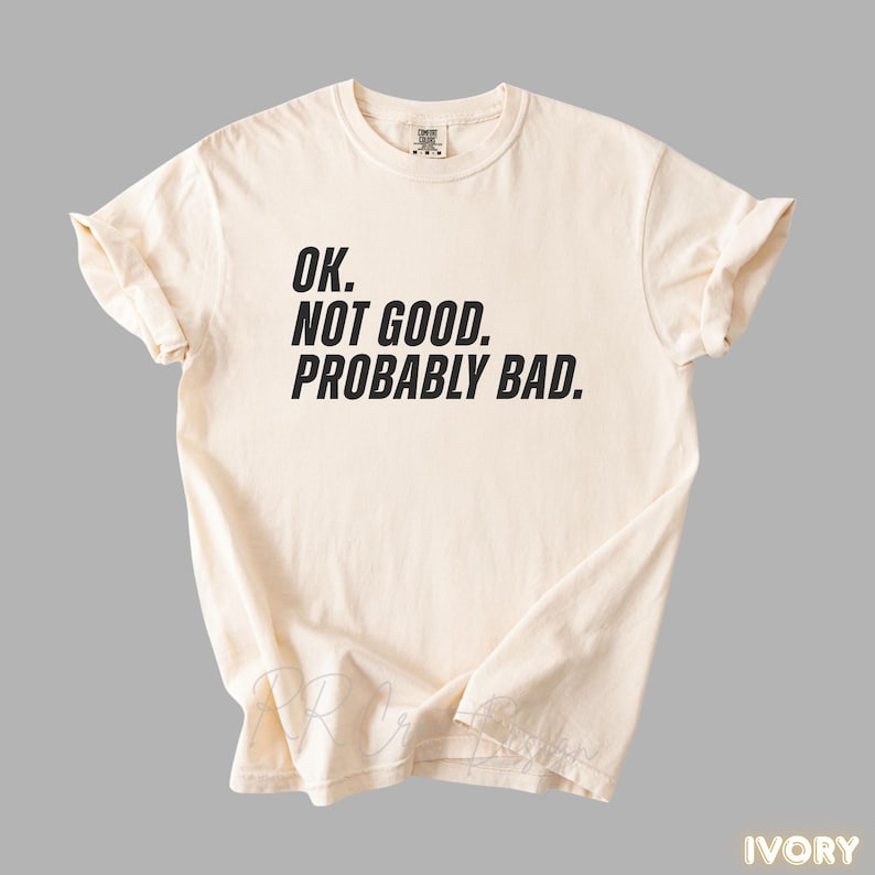 Pu&ograve; includere: T-shirt color avorio con la scritta "OK. NOT GOOD. PROBABLY BAD." in grassetto nero. La maglietta &egrave; piegata e appoggiata piatta, mostrando il design. La maglietta &egrave; realizzata in un materiale morbido.