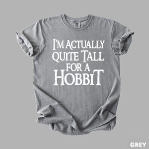 Puede incluir: Una camiseta gris con el texto blanco "I'M ACTUALLY QUITE TALL FOR A HOBBIT". La camiseta est&aacute; doblada y tiene las mangas enrolladas. La palabra "GREY" est&aacute; en la esquina inferior derecha.