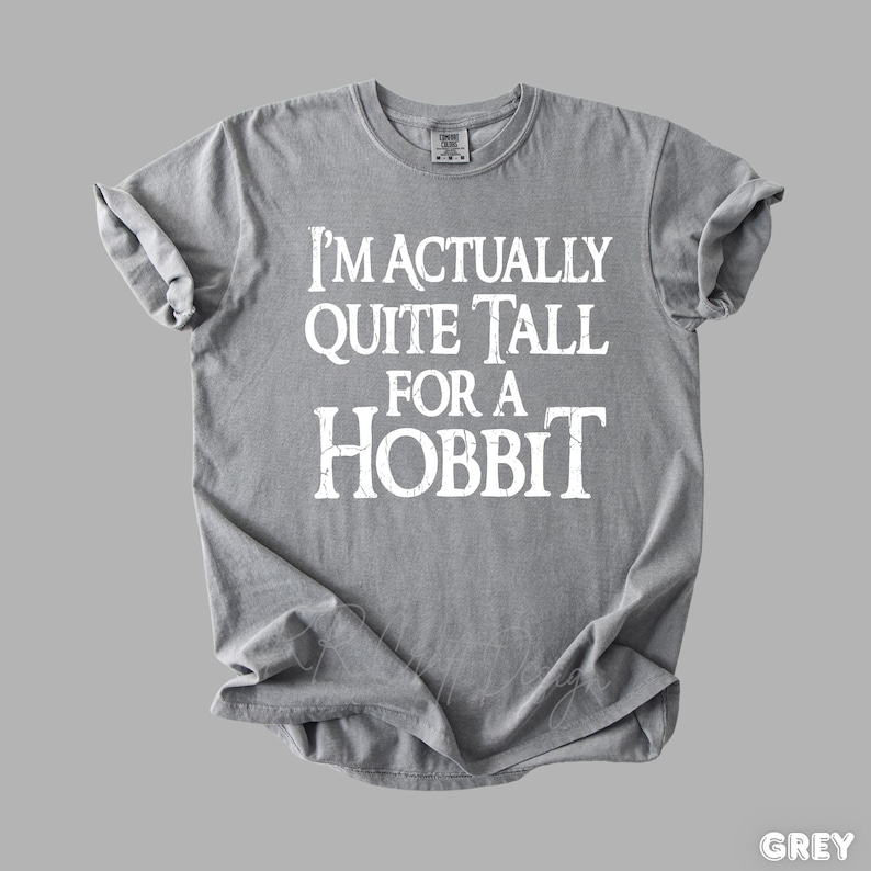 Puede incluir: Una camiseta gris con el texto blanco "I'M ACTUALLY QUITE TALL FOR A HOBBIT". La camiseta est&aacute; doblada y tiene las mangas enrolladas. La palabra "GREY" est&aacute; en la esquina inferior derecha.