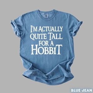 Puede incluir: Una camiseta azul claro con el texto blanco "I'M ACTUALLY QUITE TALL FOR A HOBBIT". La camiseta tiene las mangas enrolladas. El texto est&aacute; centrado en la parte delantera de la camiseta.