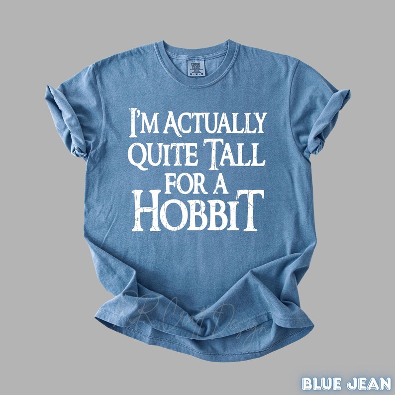 Puede incluir: Una camiseta azul claro con el texto blanco "I'M ACTUALLY QUITE TALL FOR A HOBBIT". La camiseta tiene las mangas enrolladas. El texto est&aacute; centrado en la parte delantera de la camiseta.