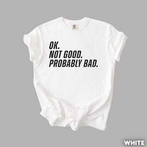 Pu&ograve; includere: Una maglietta bianca con la scritta "OK. NOT GOOD. PROBABLY BAD." stampata in nero. La maglietta ha maniche corte e girocollo. La maglietta &egrave; realizzata in un materiale morbido e viene presentata su sfondo grigio.