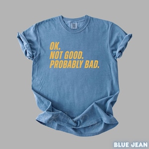 Pu&ograve; includere: Una maglietta blu jeans con la scritta "OK. NOT GOOD. PROBABLY BAD." in giallo. La maglietta &egrave; piegata e appoggiata su una superficie grigia. Le parole "BLUE JEAN" sono in basso a destra.