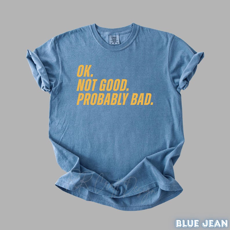 Pu&ograve; includere: Una maglietta blu jeans con la scritta "OK. NOT GOOD. PROBABLY BAD." in giallo. La maglietta &egrave; piegata e appoggiata su una superficie grigia. Le parole "BLUE JEAN" sono in basso a destra.