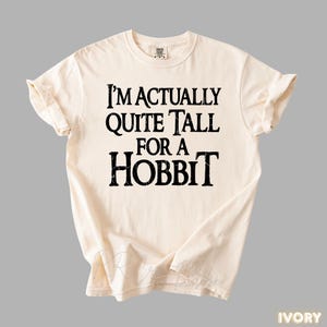 Puede incluir: Camiseta de color marfil con el texto "I'M ACTUALLY QUITE TALL FOR A HOBBIT" en letras negras y en negrita. La camiseta est&aacute; doblada y colocada plana, mostrando el dise&ntilde;o. Es una prenda c&oacute;moda e informal.