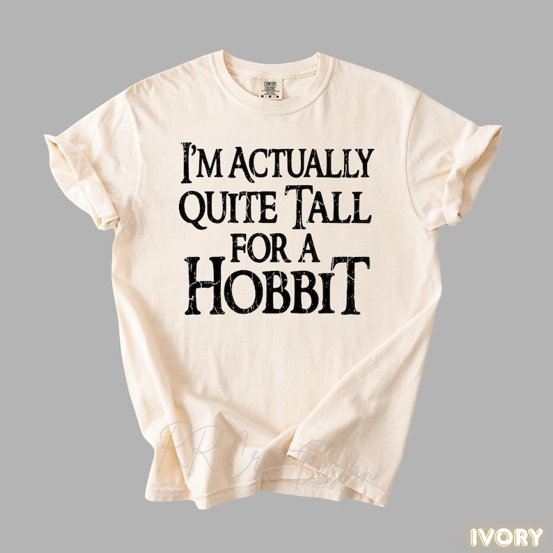 Puede incluir: Camiseta de color marfil con el texto "I'M ACTUALLY QUITE TALL FOR A HOBBIT" en letras negras y en negrita. La camiseta est&aacute; doblada y colocada plana, mostrando el dise&ntilde;o. Es una prenda c&oacute;moda e informal.