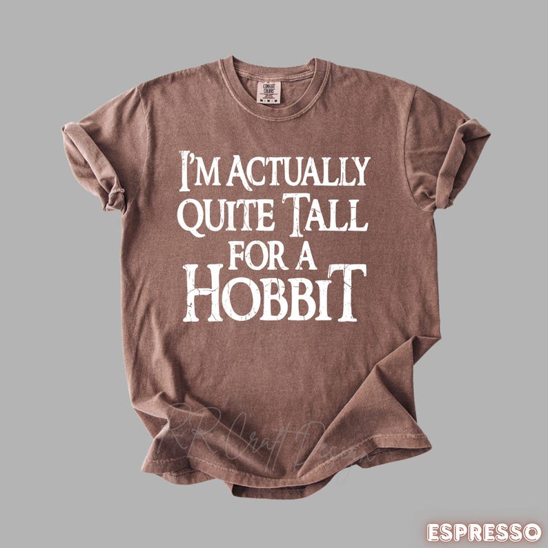 Puede incluir: Camiseta color espresso con el texto blanco "I'M ACTUALLY QUITE TALL FOR A HOBBIT". La camiseta tiene cuello redondo y mangas cortas. La tela parece suave y ligeramente arrugada.