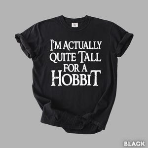 Puede incluir: Camiseta negra con el texto blanco "I'M ACTUALLY QUITE TALL FOR A HOBBIT". La camiseta tiene mangas cortas y cuello redondo. La camiseta est&aacute; hecha de un material suave.