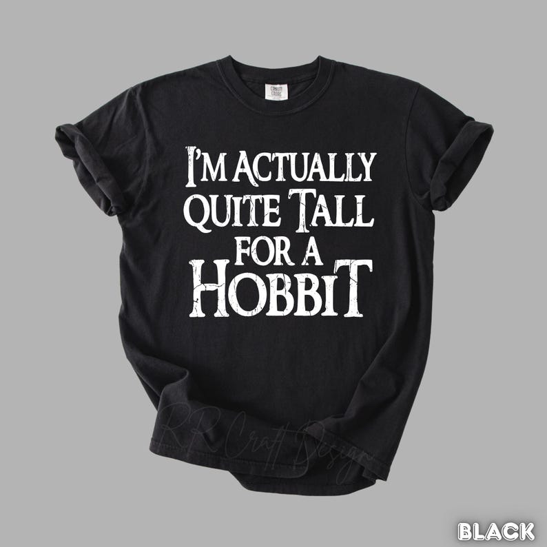 Puede incluir: Camiseta negra con el texto blanco "I'M ACTUALLY QUITE TALL FOR A HOBBIT". La camiseta tiene mangas cortas y cuello redondo. La camiseta est&aacute; hecha de un material suave.