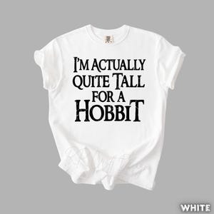 Puede incluir: Camiseta blanca con la frase "I'M ACTUALLY QUITE TALL FOR A HOBBIT" en negro, con un estilo desgastado. La camiseta es de cuello redondo y parece estar hecha de un material suave. El tema general es la literatura fant&aacute;stica.