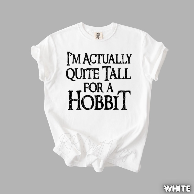 Puede incluir: Camiseta blanca con la frase "I'M ACTUALLY QUITE TALL FOR A HOBBIT" en negro, con un estilo desgastado. La camiseta es de cuello redondo y parece estar hecha de un material suave. El tema general es la literatura fant&aacute;stica.