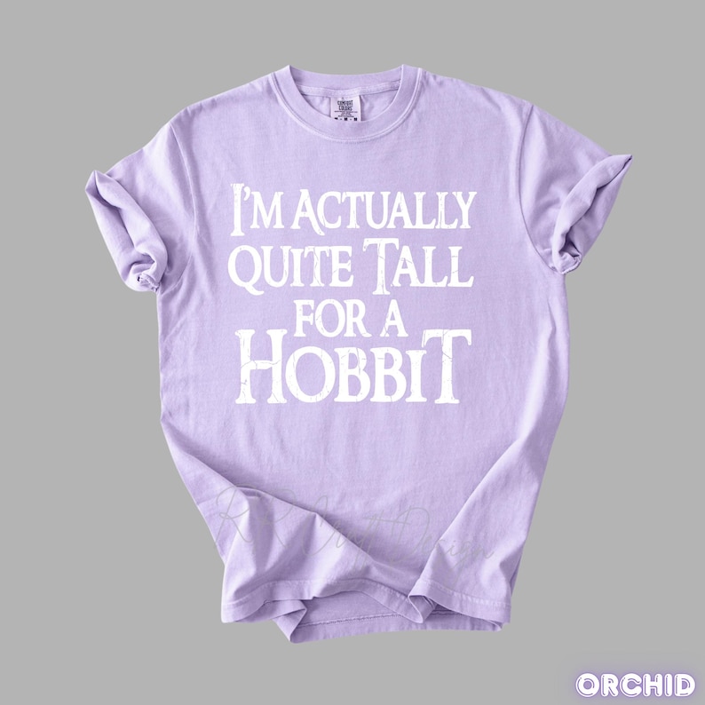 Puede incluir: Una camiseta de color lila claro con el texto blanco "I'M ACTUALLY QUITE TALL FOR A HOBBIT". Las mangas est&aacute;n enrolladas. La camiseta est&aacute; sobre un fondo gris. Es una prenda informal.