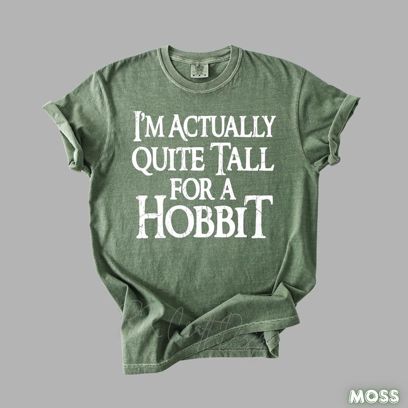 Puede incluir: Una camiseta verde musgo con el texto blanco "I'M ACTUALLY QUITE TALL FOR A HOBBIT". La camiseta tiene cuello redondo y mangas cortas. La palabra "MOSS" est&aacute; en blanco en la esquina inferior derecha.