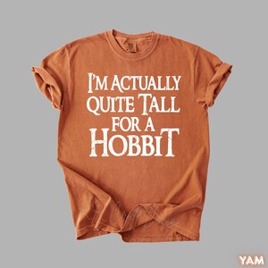 Puede incluir: Camiseta de color naranja quemado con el texto "I'M ACTUALLY QUITE TALL FOR A HOBBIT" en blanco. La camiseta tiene cuello redondo y mangas cortas. La tela parece suave y ligeramente descolorida.