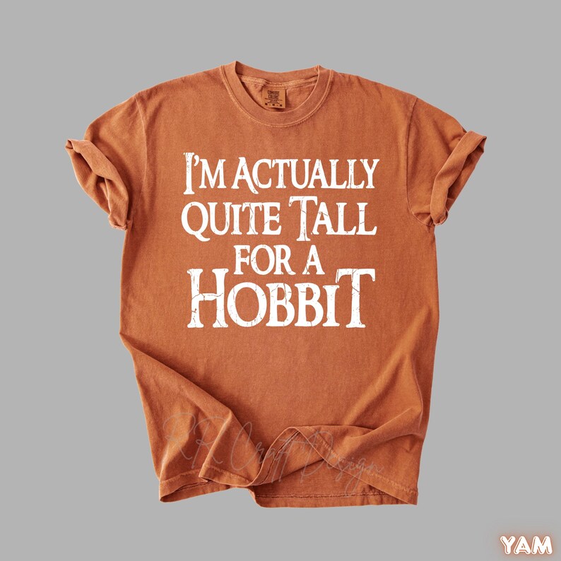 Puede incluir: Camiseta de color naranja quemado con el texto "I'M ACTUALLY QUITE TALL FOR A HOBBIT" en blanco. La camiseta tiene cuello redondo y mangas cortas. La tela parece suave y ligeramente descolorida.