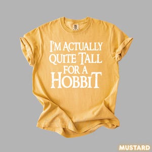 Puede incluir: Camiseta de color mostaza con el texto blanco "I'M ACTUALLY QUITE TALL FOR A HOBBIT". La camiseta est&aacute; doblada y tiene cuello redondo. La palabra "MUSTARD" est&aacute; en la esquina inferior derecha.