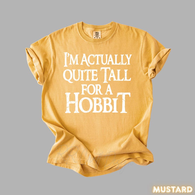 Puede incluir: Camiseta de color mostaza con el texto blanco "I'M ACTUALLY QUITE TALL FOR A HOBBIT". La camiseta est&aacute; doblada y tiene cuello redondo. La palabra "MUSTARD" est&aacute; en la esquina inferior derecha.