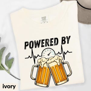 May include: Ivory-colored t-shirt with the words "POWERED BY" above a beer-themed graphic. The graphic includes a heartbeat line, a gauge, and two beer mugs with foam. The shirt is a Comfort Colors brand.