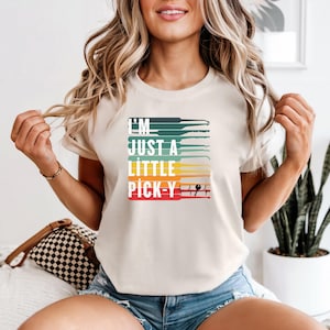 May include: Cream-colored t-shirt with the text "I'M JUST A LITTLE PICK-Y" in a colorful, retro design. The graphic includes lock picks in shades of green, blue, yellow, and red. A fun and unique tee for those who appreciate locksmithing or security.