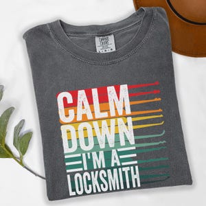 May include: Dark gray t-shirt with the text "CALM DOWN I'M A LOCKSMITH" in white, red, orange, yellow, green, and blue. The shirt has a crew neck and a Comfort Colors tag.