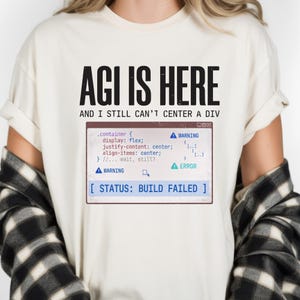 May include: A cream-colored t-shirt featuring the black text AGI IS HERE AND I STILL CAN'T CENTER A DIV above a graphic of a computer window displaying CSS code, warning icons, and the blue text STATUS: BUILD FAILED in a rectangular box.