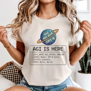 May include: A cream-colored short-sleeve t-shirt featuring a graphic of a blue planet with a gold ring. The text reads: THE AGI AGE GPT-6. Below, a box contains: AGI IS HERE, [SYS_INFO] AGI_STATUS: ARRIVED, [CORE] Model: GPT-6 Astra, [STATE] Complete., [MSG] Hello, World.