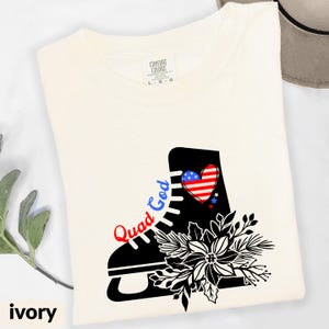 May include: Ivory-colored t-shirt featuring a black ice skate graphic with a red, white, and blue heart and stars. The words "Quad God" are written in red and blue. A floral design is below the skate.