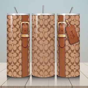 May include: A cylindrical tumbler with a straw, featuring a tan and brown repeating logo pattern. The design includes a vertical brown strap graphic with a gold-toned buckle labeled COACH and a hanging brown leather-style luggage tag with a logo.