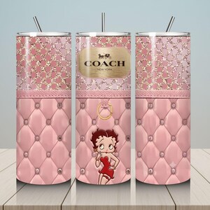May include: A pink tumbler with a glittery top and a quilted bottom. The tumbler features a Betty Boop graphic and a gold Coach New York logo. Includes a silver straw.