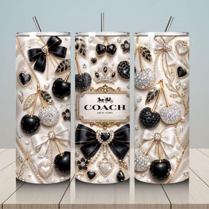May include: A cylindrical tumbler with a straw featuring a cream-colored satin background decorated with black and crystal-encrusted cherries, bows, hearts, and pearls. The center displays a gold-framed plaque with the text COACH NEW YORK.