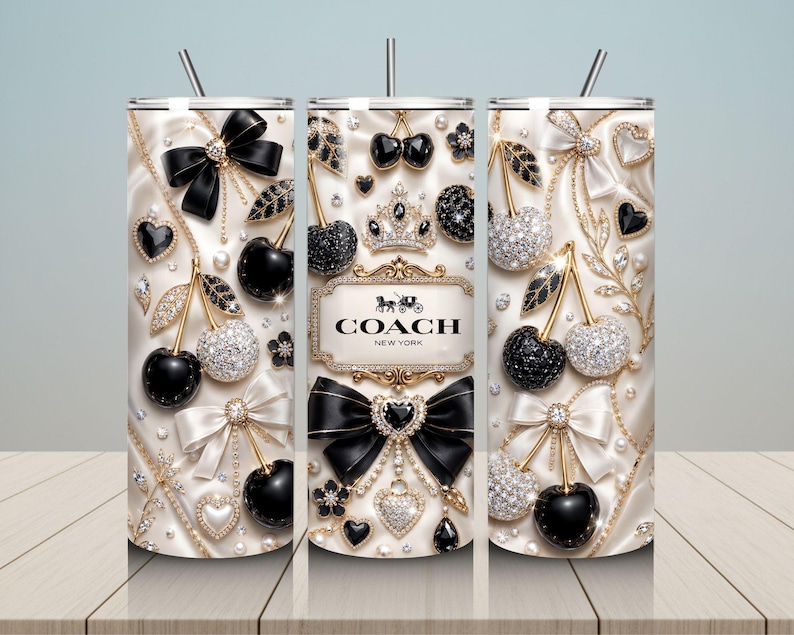 May include: A cylindrical tumbler with a straw featuring a cream-colored satin background decorated with black and crystal-encrusted cherries, bows, hearts, and pearls. The center displays a gold-framed plaque with the text COACH NEW YORK.