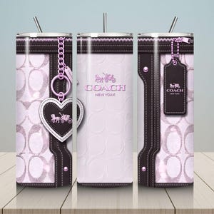 May include: A cylindrical tumbler featuring a light pink and dark brown design with a repeating logo pattern. The exterior displays a zipper graphic, a heart-shaped charm, and a rectangular luggage tag, all printed with the Coach New York logo.