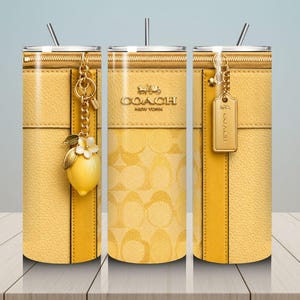 May include: Yellow Coach tumbler with a zipper design, gold accents, and a lemon charm. The tumbler features the Coach New York logo and a straw. The design includes a textured leather-like appearance and a repeating pattern.