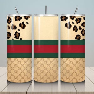 May include: Three cylindrical tumblers with lids and straws. The design features a top section with a black and gold leopard print, a middle section with a horizontal red stripe bordered by two green stripes, and a bottom section with a gold monogram pattern.