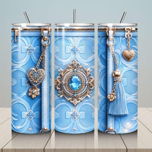 May include: A light blue tumbler with a decorative design. The tumbler features a zipper top, a heart-shaped charm, and a tassel. The design includes a floral pattern and a large, ornate jewel in the center. A straw is inserted in the lid.