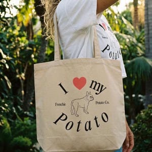 Könnte beinhalten: Beige Tragetasche mit dem Text "I ❤️ my Potato" und einer Grafik einer Französischen Bulldogge. Die Tasche enthält auch den Text "Frenchie Potato Co." in Schwarz. Die Tasche wird von einer Person getragen.