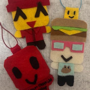 May include: Three handmade felt ornaments featuring blocky character designs. One is a yellow figure in a red shirt, another is a red square with a smiling face and text, and the third is a figure wearing a burger hat and light blue glasses.