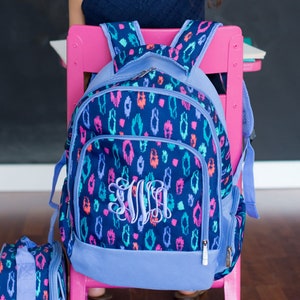 May include: A navy blue backpack featuring a vibrant, multi-colored animal print pattern in shades of pink, purple, and teal. The bag includes a front zippered pocket with a large, light blue embroidered monogram and adjustable shoulder straps.