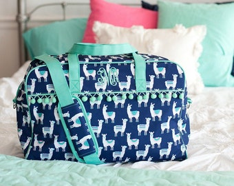 May include: A navy blue duffel bag featuring a repeating white and light blue llama pattern, mint green trim, and a row of mint green pom-poms. The bag includes a shoulder strap and a white monogrammed letter detail on the front exterior panel.