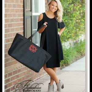 May include: A black tote bag with a red monogram on the front. The bag is being held by a person wearing a black dress and grey ankle boots.