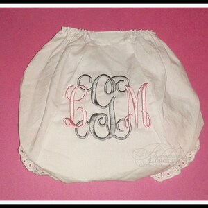 May include: White cotton baby bloomers with a pink and grey monogram "LGM" embroidered on the front. The bloomers have a white lace trim around the edges.