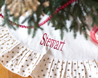May include: A cream-colored Christmas tree skirt featuring the name Stewart embroidered in red thread. The skirt has a pleated border decorated with a metallic gold polka dot pattern, positioned beneath the branches of a pine tree.
