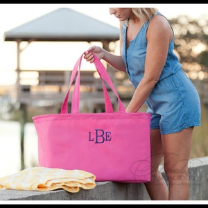 May include: A pink tote bag with a blue monogram "LBE" embroidered on the front. The bag is large enough to hold a beach towel and other items.