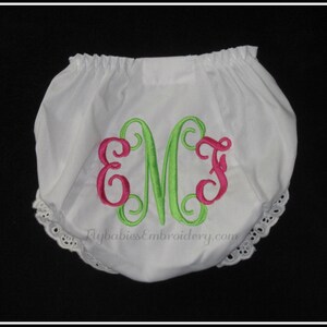 May include: White baby bloomers with a green and pink monogram embroidered on the front. The bloomers have a lace trim around the leg openings. The text "FlybabiesEmbroidery.com" is printed on the front.