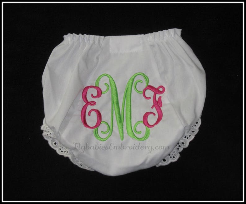 May include: White baby bloomers with a green and pink monogram embroidered on the front. The bloomers have a lace trim around the leg openings. The text "FlybabiesEmbroidery.com" is printed on the front.