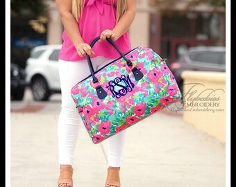 May include: A large rectangular duffel bag featuring a vibrant floral pattern in shades of hot pink, teal, and white. The bag has dark navy blue handles and a dark navy blue embroidered monogram with the letters RSM in the center.